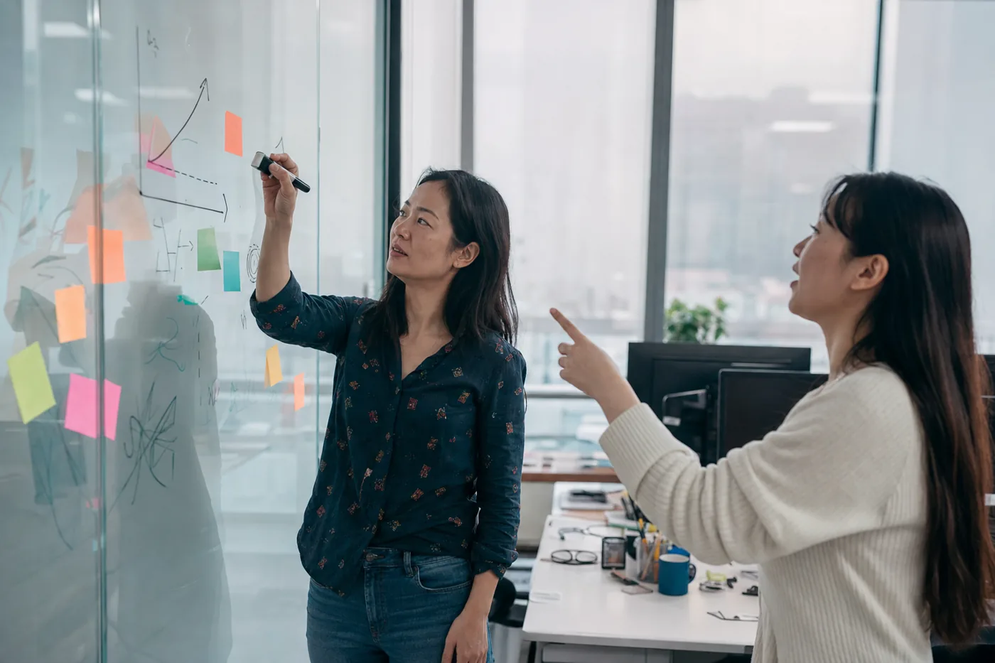 A person drawing a rising learning curve and sticky notes on a glass office wall while a colleague reacts
