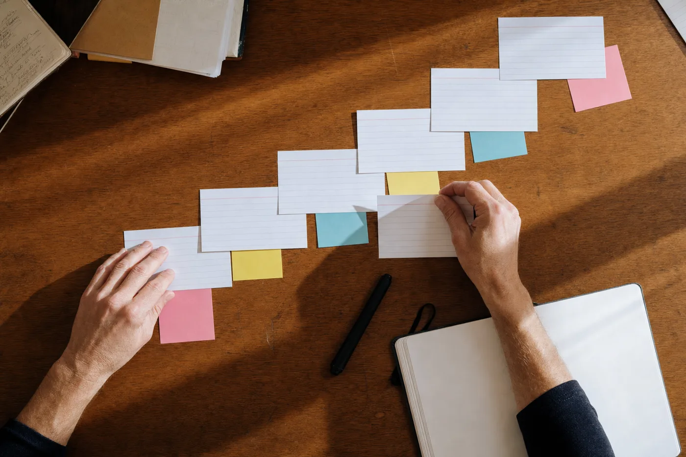 Overhead view of hands arranging sticky notes and index cards into a rising curve on a wooden desk, representing employee skills improving through repeated practice