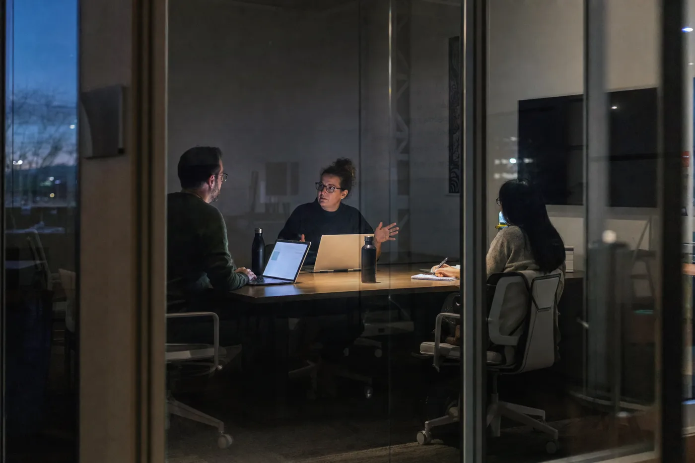 Two employees rehearsing a difficult scenario in a glass-walled meeting room while a colleague observes and takes notes