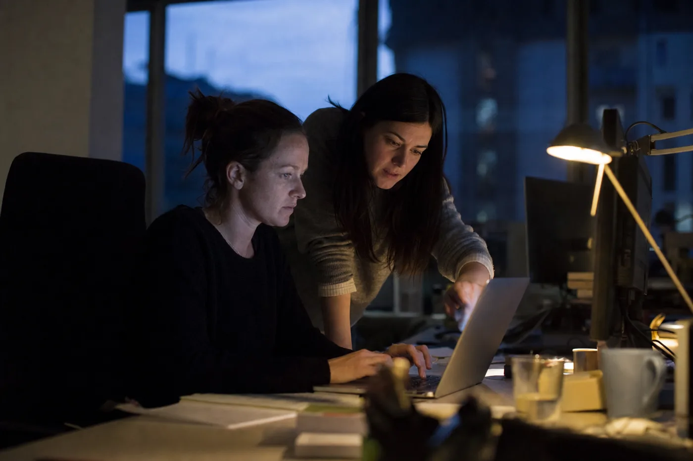 Two colleagues practicing an AI-assisted task together at a laptop in the evening, one giving feedback