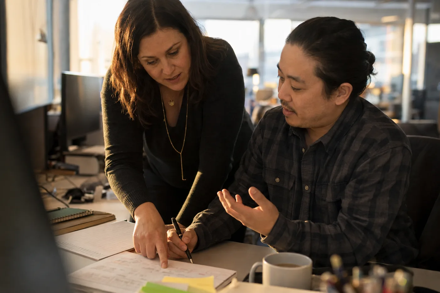 A trainer leaning over a colleague's desk, pointing at a worksheet while the colleague talks through their reasoning, warm afternoon window light