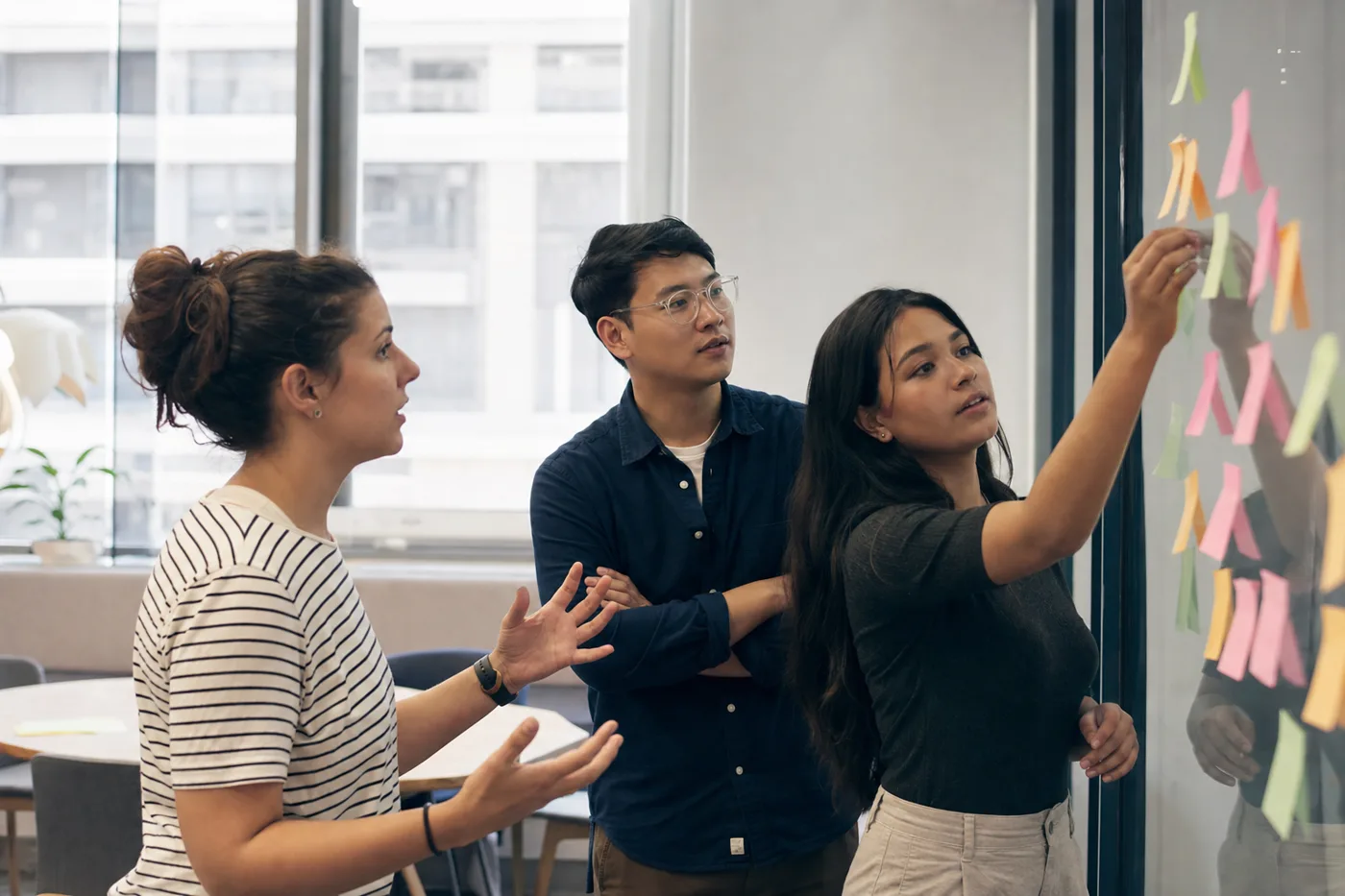 Three new hires gathered at a glass wall covered in sticky notes during an onboarding session, one mid-sentence explaining an idea, bright overcast daylight
