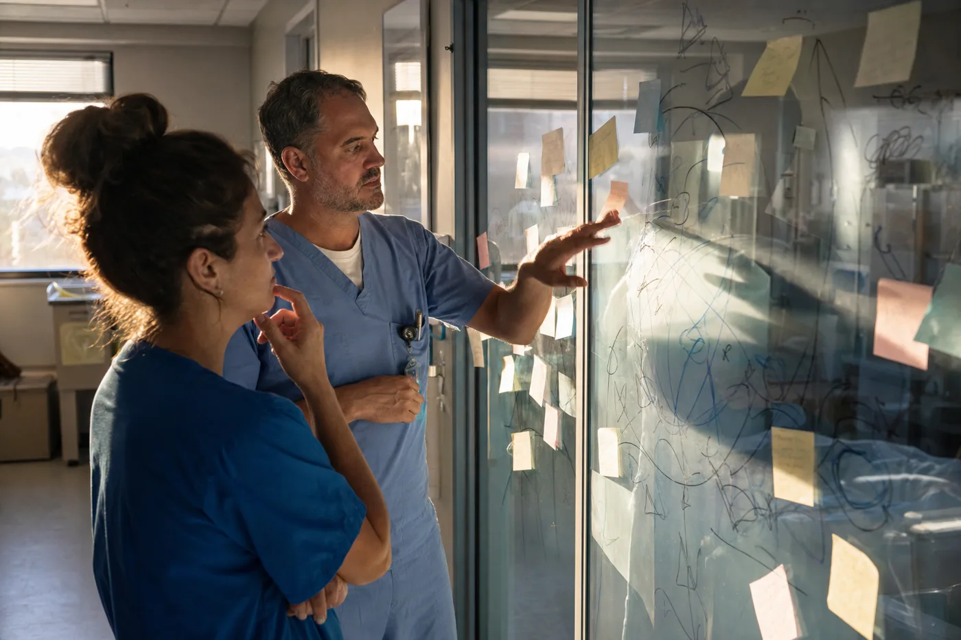 Two hospital staff working through hand-drawn charts and sticky notes on a glass wall, planning how to measure frontline training outcomes