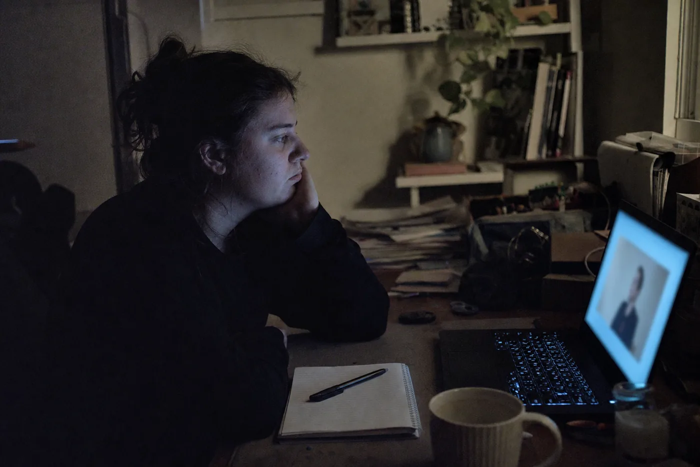 An employee slumped at a desk, half-watching a dull training module glow on a laptop while real work sits untouched beside them, moody evening office light with visible grain