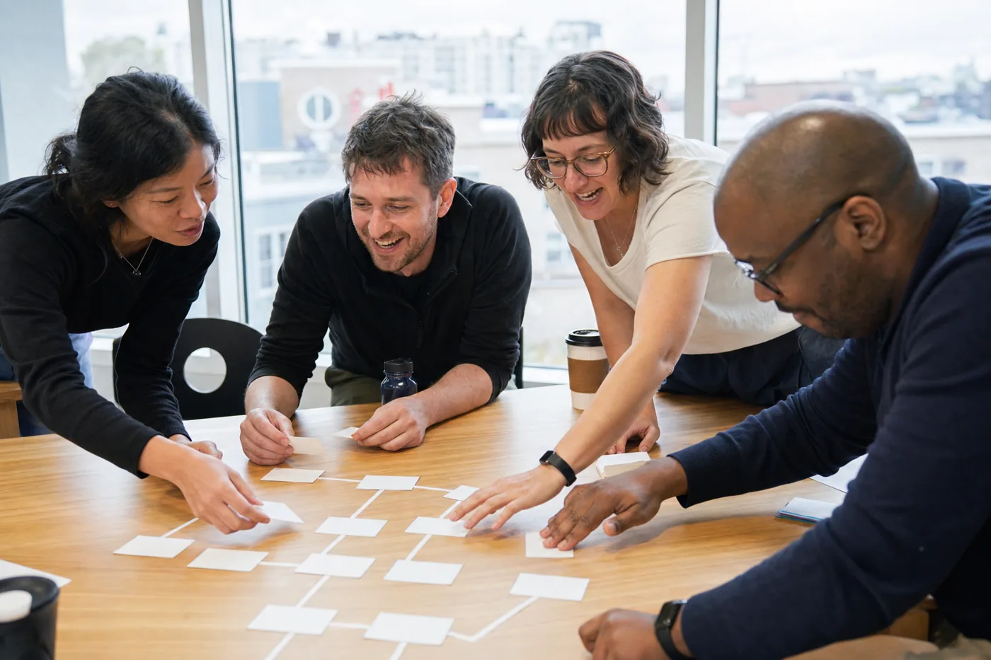 Colleagues leaning in around a table, arranging printed scenario cards into a decision path during a hands-on training workshop, bright overcast daylight through tall windows