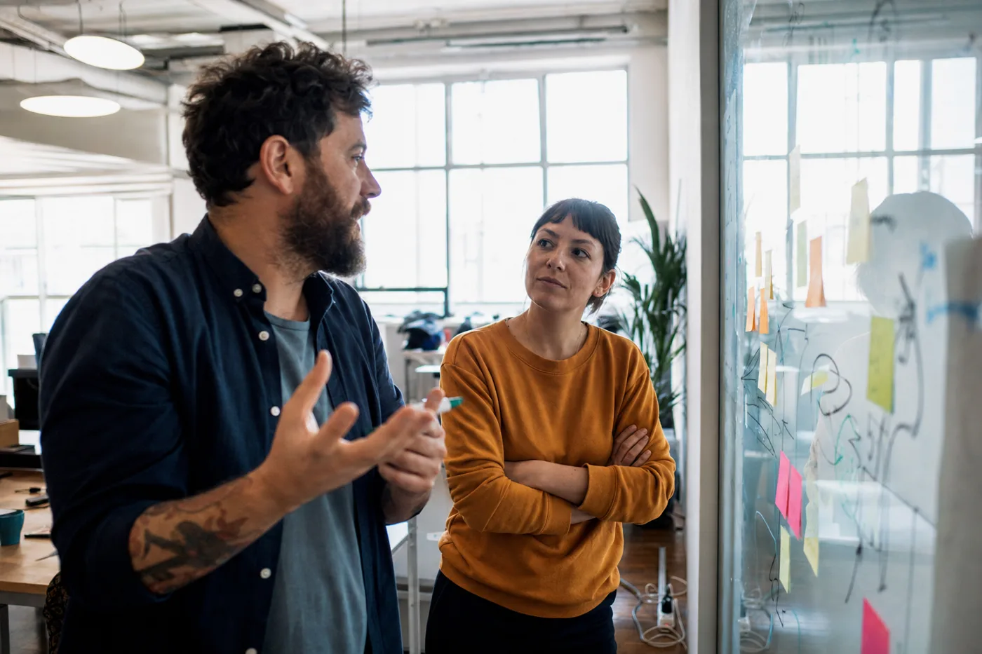 Two coworkers standing at a glass wall covered in hand-drawn arrows and sticky notes, working a problem out together as an example of productive struggle