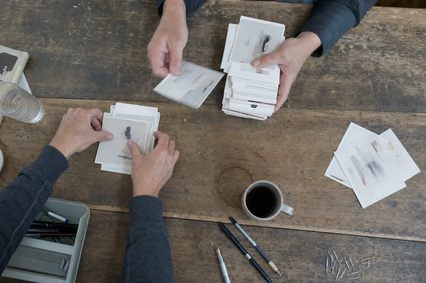 Overhead view of two colleagues' hands sorting a tall stack of printed cards into one small pile on a wooden desk
