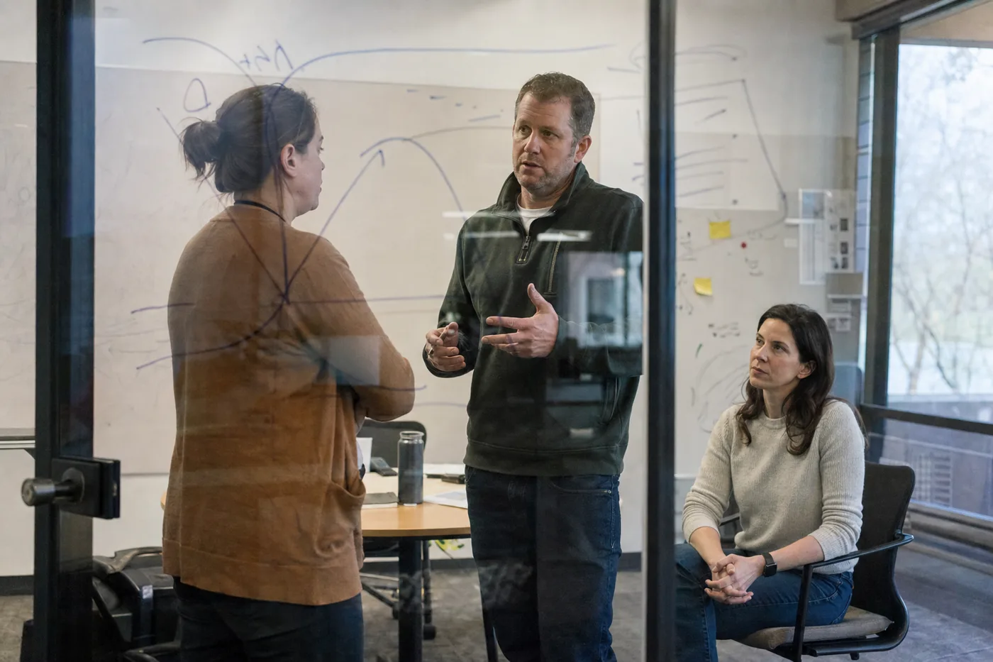 Two coworkers rehearsing a difficult customer conversation face to face in a glass-walled room while a colleague observes, practicing the skill rather than watching a video