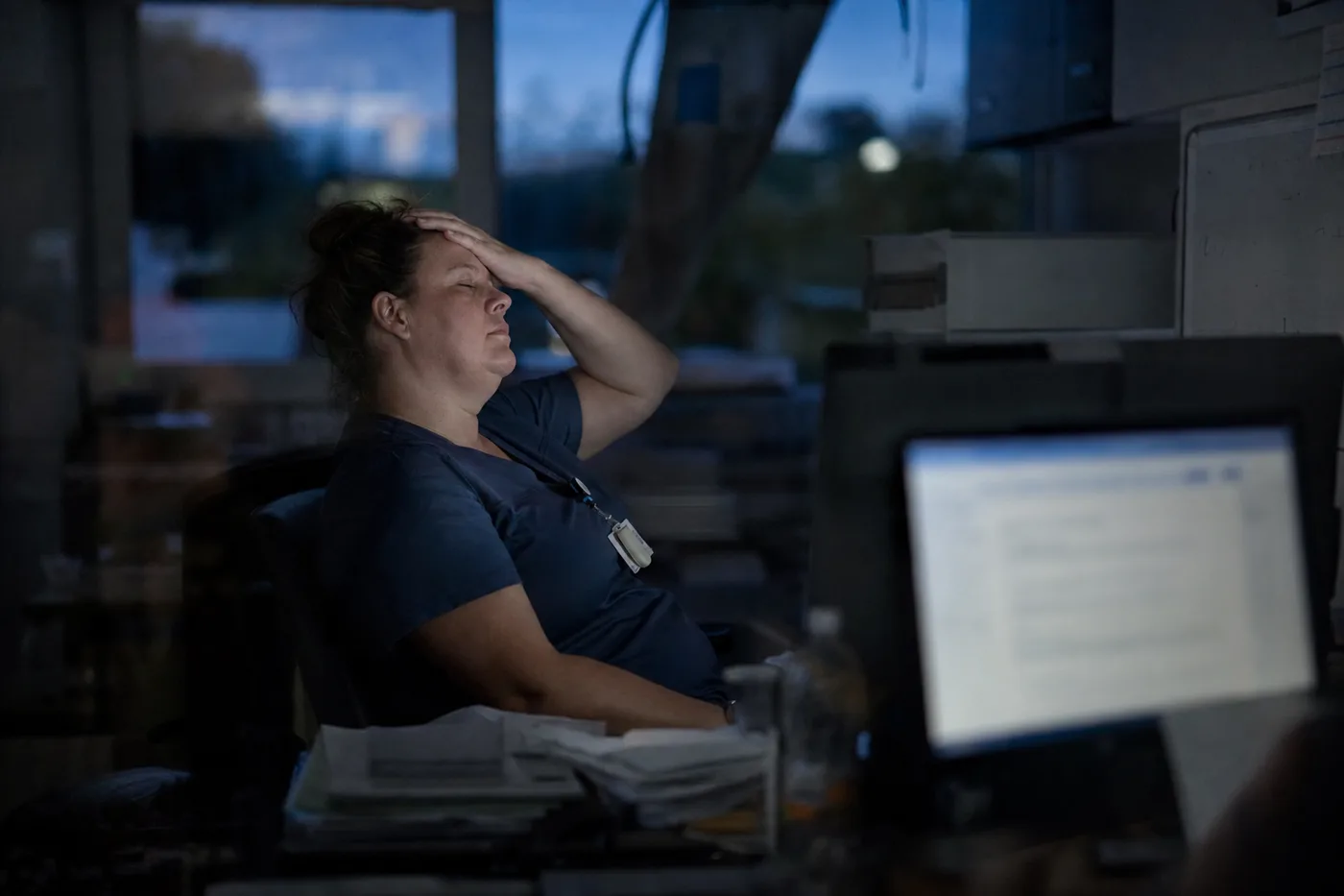 A clinician pauses at dusk beside a blurred monitor with her hand on her forehead, after an AI-assisted shift when unassisted skills still matter